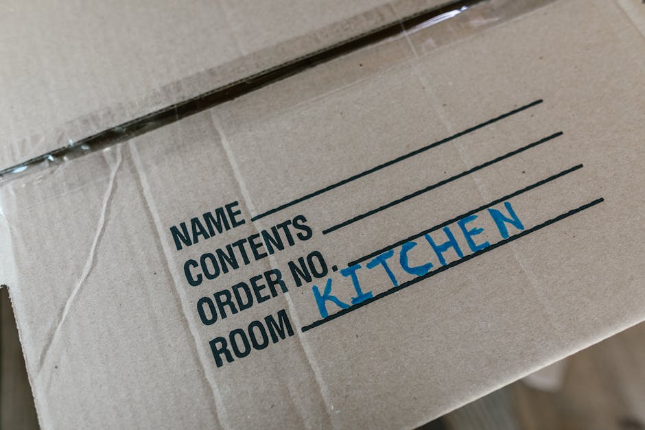 Close-up image of a cardboard moving box with a label indicating it belongs to the kitchen, used in a home relocation process. The box is sealed with clear packing tape, and the label features printed