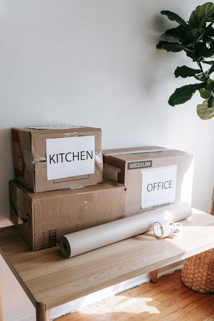 A close-up view of several packed cardboard boxes placed on a light wooden table, preparing for a home relocation or office move, with labels indicating their contents, including one labeled 'KITCHEN'