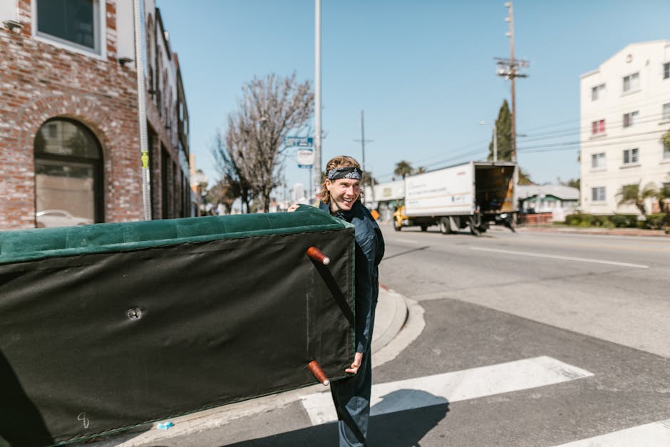 Two movers from Man and Van Merton are loading a large, dark green upholstered furniture item into the rear of a white van parked on a suburban street. The furniture is partially inside the vehicle, w